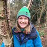 Alice wearing green hat and blue coat and standing next to a tree