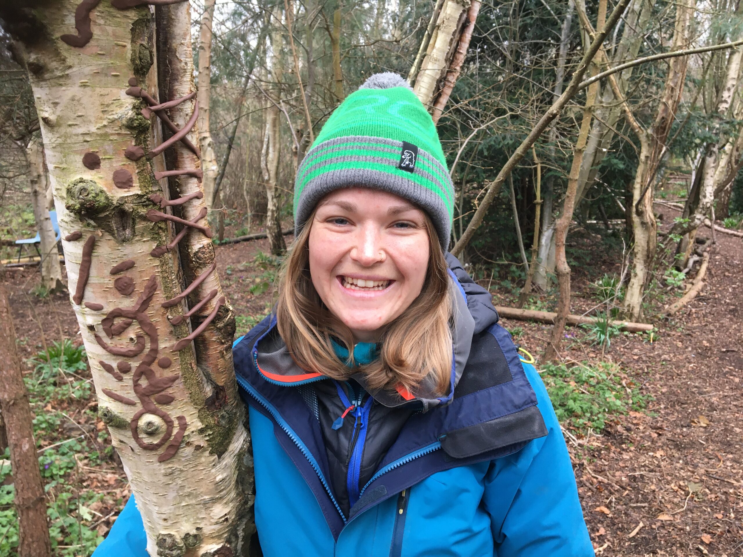 Alice wearing green hat and blue coat and standing next to a tree