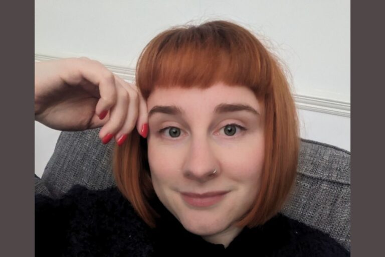 Hannah has red hair in a bob and is leaning against her hand slightly smiling at the camera.