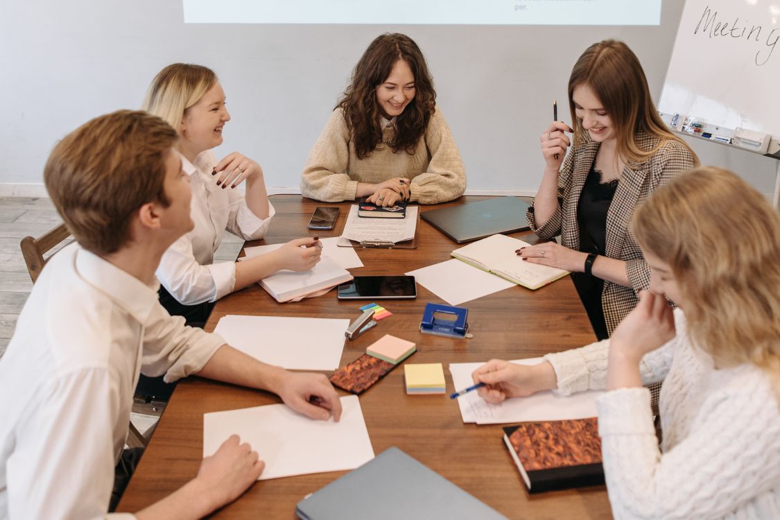 Group of people sat around a wooden meeting table in a meeting. Everyone looks happy.