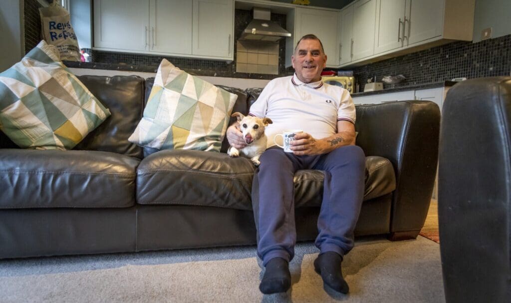 Man sat on brown sofa with small dog, holding a cuppa.