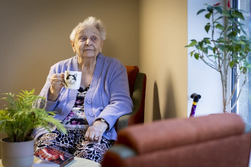 Lady in lilac cardigan drinking cuppa, the cup has a cat on it.