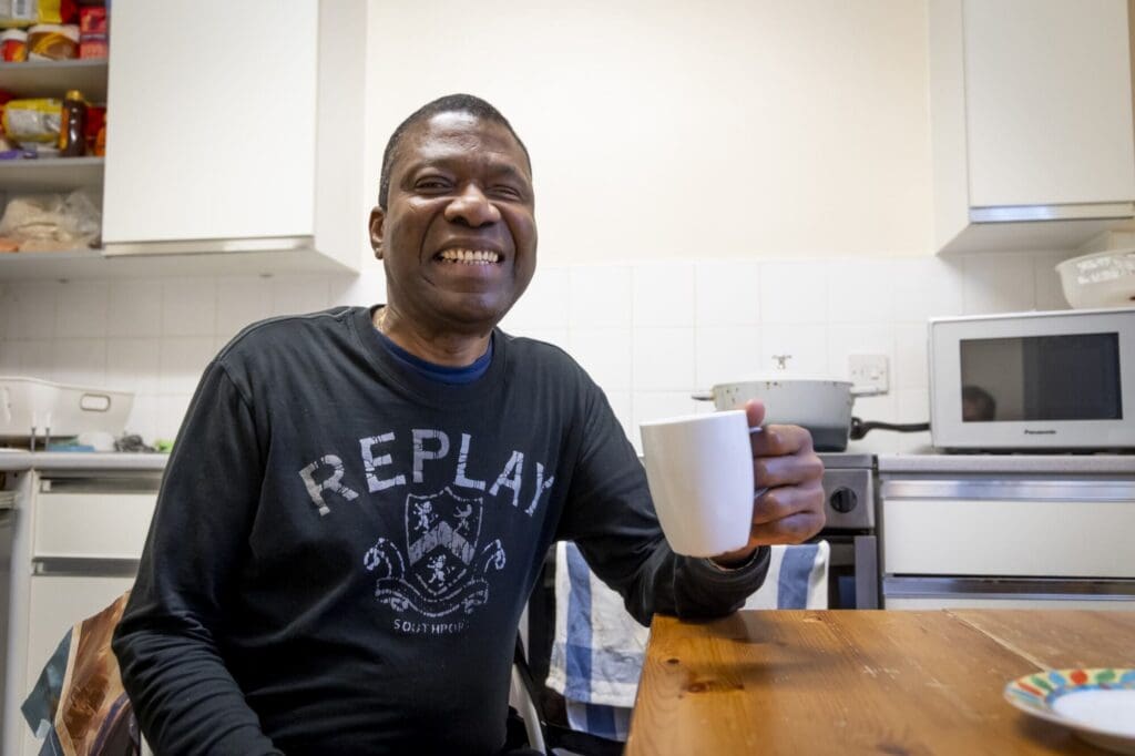 Smiling man sat at kitchen table holding a cup. Wearing a black replay top.