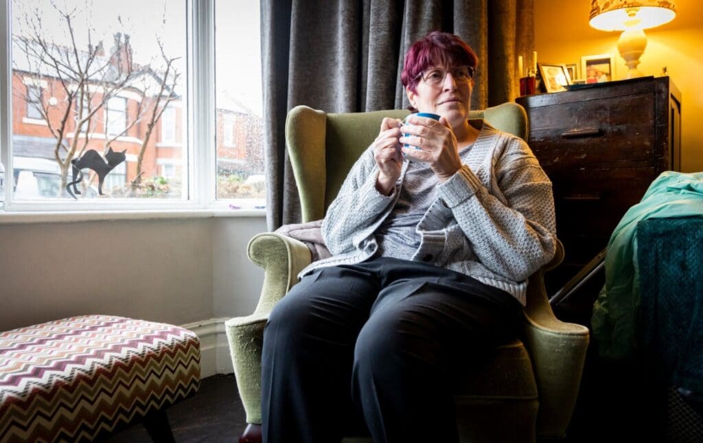 Woman sat in green armchair next to the window, holding a cup.