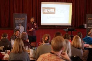 Presenter at MHA event. Standing in front of audience, wearing red.