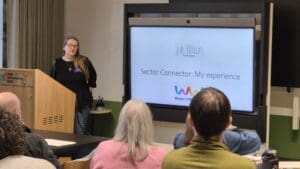 Close up shot of woman presenting. The presentation screen says 'Sector Connector - My Experience'