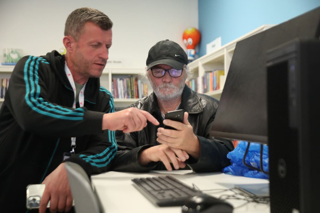 Volunteer Jim in black hoodie with blue stripes on arms is sat next to a man in a cap with a white beard and glasses in a black jacket. He is holding a phone and Jim is pointing to the phone.