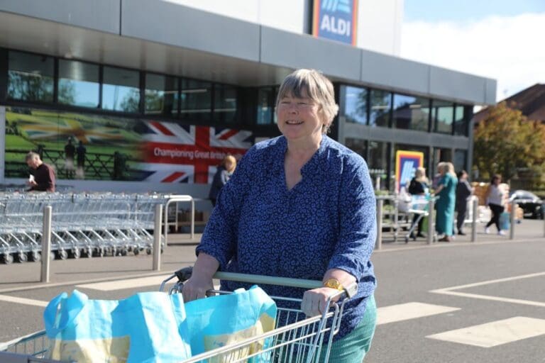 Woman in blue top pushing a shopping trolley full of bags in front of a supermarket