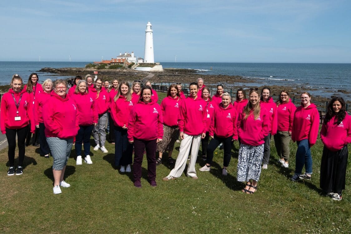 Team of people in pink hoodies pose by the sea, St Mary's Lighthouse in background.