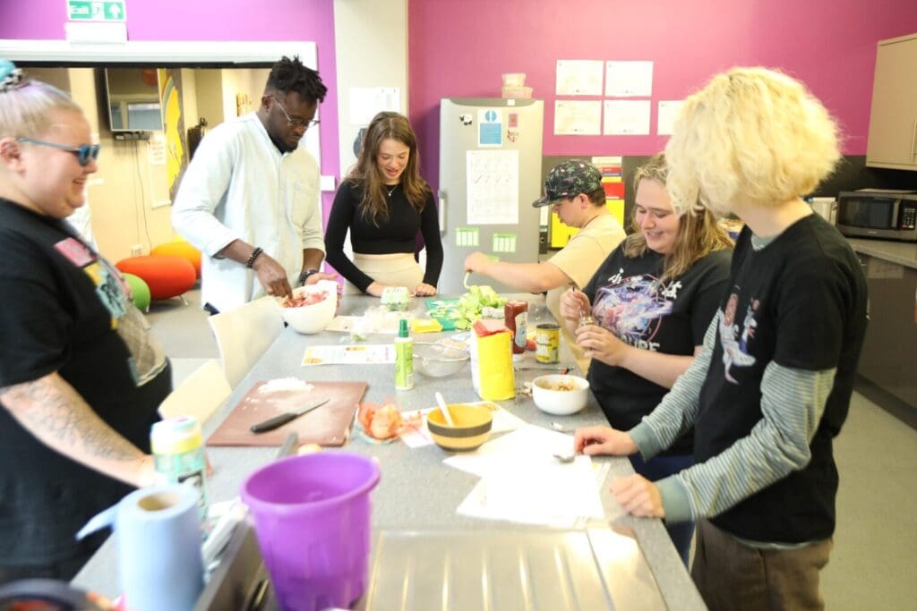 A group of people gathered in a kitchen, interacting and preparing food at a table. The setting includes various tableware and features an indoor atmosphere with walls visible around them.