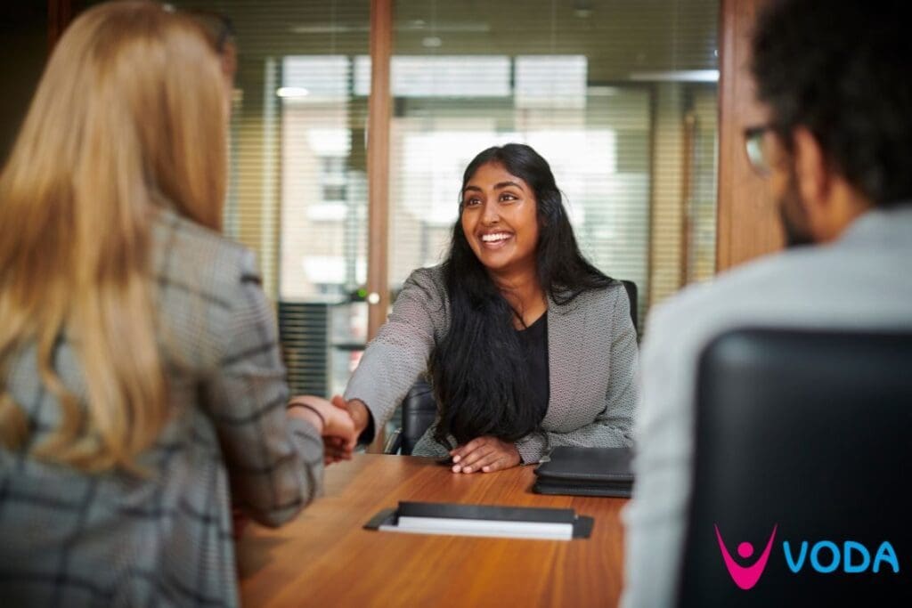 Two women is smart business wear shake hands over a table in an indoor office setting, smiling at each other amidst the conversation. You can also see the back of another person with short dark hair sat at the table.