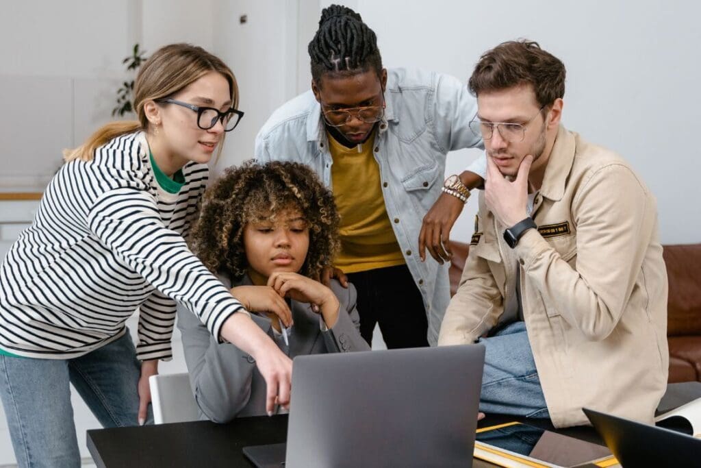 A group of people sitting indoors, gathered around a laptop, engaged in discussion and looking at the screen. The setting includes a wall in the background, and various individuals are wearing different clothing styles while focused on the computer.